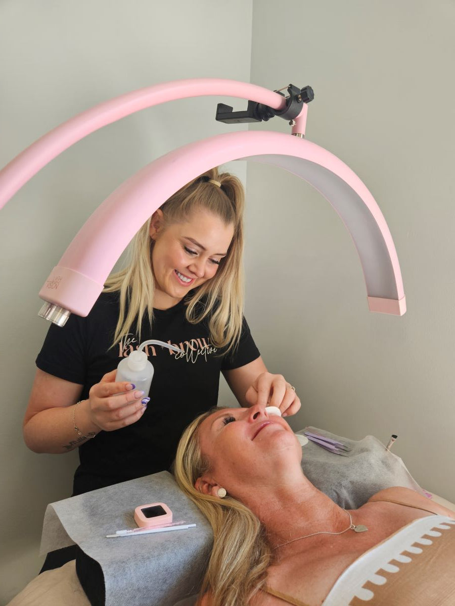 Lash artist performing a professional lash lift treatment under a pink LED lamp.